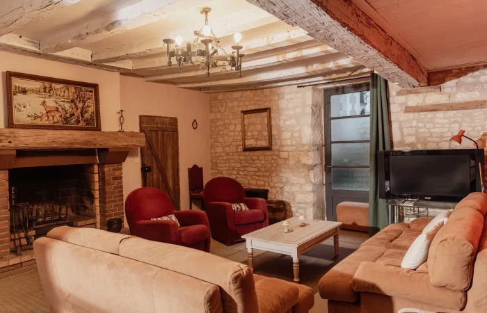 Grand salon du gîte Le Presbytère au Parc de Lesseps — cheminée en brique, murs en pierre, canapés caramel et fauteuils bordeaux, poutres apparentes, hébergement de caractère en Berry Indre